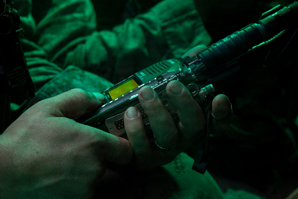 A close-up image showing two hands holding a tactical military radio or communication device, illuminated by green light. The scene appears to be a low-light or nighttime operational environment, with military uniform fabric visible in the background. Additional equipment, including what appears to be a weapon barrel or rail system, is visible in the upper portion of the frame.
