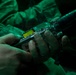 A close-up image showing two hands holding a tactical military radio or communication device, illuminated by green light. The scene appears to be a low-light or nighttime operational environment, with military uniform fabric visible in the background. Additional equipment, including what appears to be a weapon barrel or rail system, is visible in the upper portion of the frame.