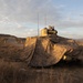 A military ground vehicle is partially concealed beneath a desert-pattern camouflage net in an arid, hilly training or operational environment. The vehicle's turret and two radio antennas are visible above the netting. The scene appears to be taken during golden hour lighting, suggesting early morning or late afternoon.