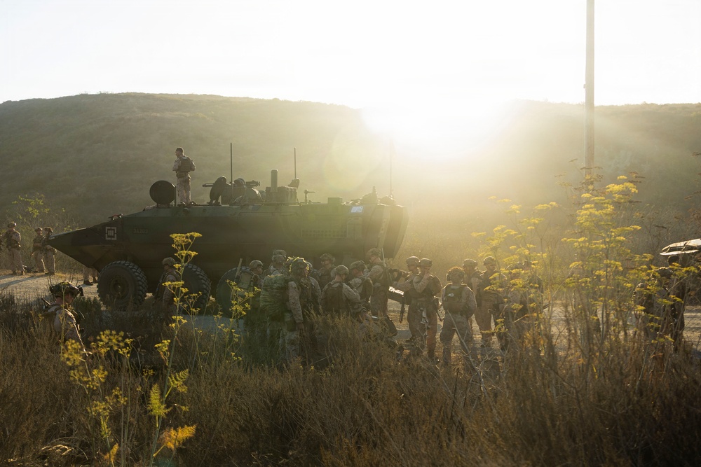 A group of military personnel in combat gear are assembled around a large armored wheeled vehicle, likely during a training exercise or operational briefing. The scene is set in a dry, hilly terrain with golden-hour sunlight creating strong backlighting. One soldier is visible standing atop the armored vehicle while others are gathered on the ground nearby.