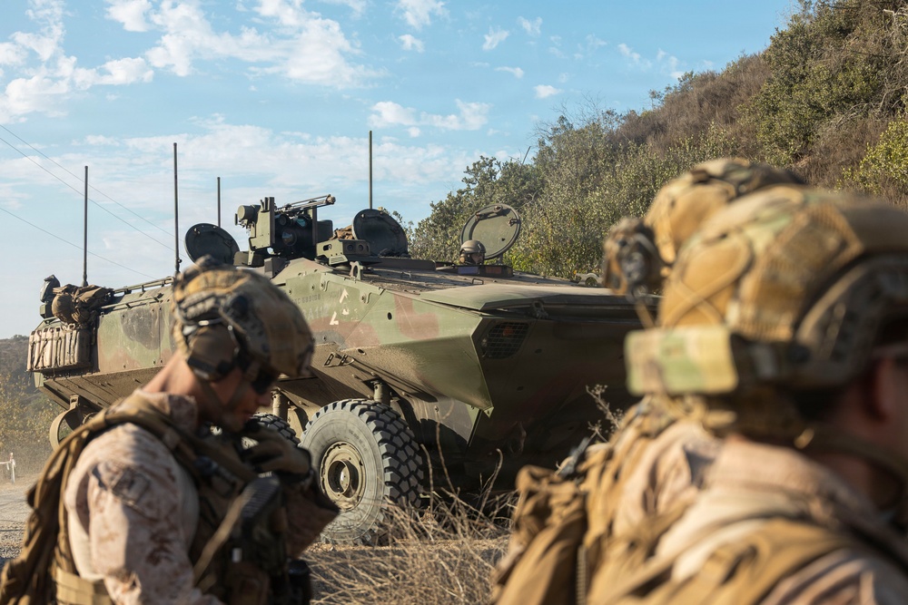 The image shows military personnel in full combat gear positioned alongside a wheeled armored fighting vehicle, likely during a training exercise or tactical operation. Two soldiers in the foreground wear camouflage uniforms and tactical helmets, while a crew member is visible in the vehicle's open hatch. The setting appears to be a dry, scrub-covered hillside terrain under a partly cloudy sky.