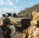 The image shows military personnel in full combat gear positioned alongside a wheeled armored fighting vehicle, likely during a training exercise or tactical operation. Two soldiers in the foreground wear camouflage uniforms and tactical helmets, while a crew member is visible in the vehicle's open hatch. The setting appears to be a dry, scrub-covered hillside terrain under a partly cloudy sky.