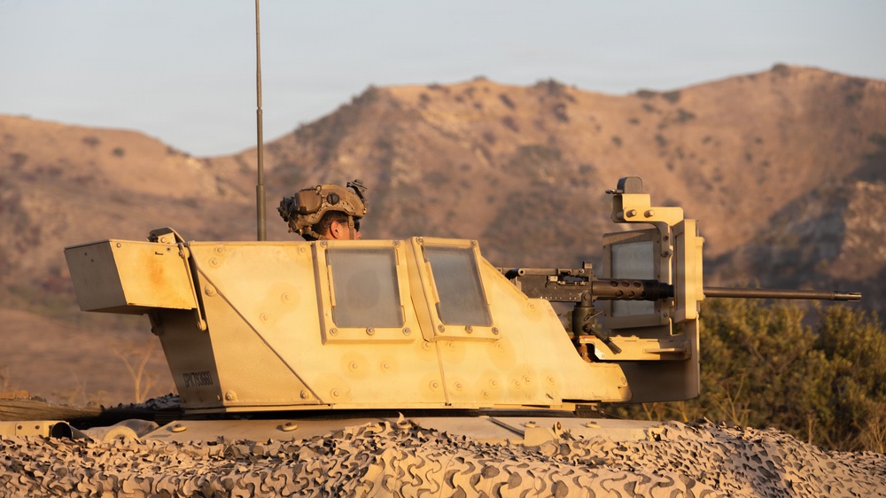 A military armored vehicle, likely a Bradley Fighting Vehicle or similar IFV, is positioned in a desert/arid terrain environment during golden hour lighting. A soldier wearing a combat helmet is visible in the commander's hatch, with a mounted weapon system (appears to be an M2 .50 caliber machine gun) visible on the right side of the turret. The vehicle is partially concealed with camouflage netting draped along its lower hull.