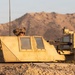 A military armored vehicle, likely a Bradley Fighting Vehicle or similar IFV, is positioned in a desert/arid terrain environment during golden hour lighting. A soldier wearing a combat helmet is visible in the commander's hatch, with a mounted weapon system (appears to be an M2 .50 caliber machine gun) visible on the right side of the turret. The vehicle is partially concealed with camouflage netting draped along its lower hull.
