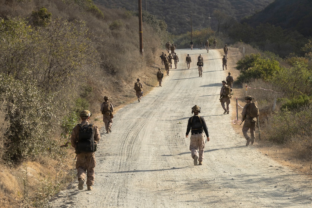 A group of military personnel in combat uniforms and gear are marching along a gravel road through dry, hilly terrain. The soldiers are spread across the road in a loose column formation, moving away from the camera. The environment appears arid with dry brush and sparse vegetation on both sides of the road.