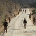 A group of military personnel in combat uniforms and gear are marching along a gravel road through dry, hilly terrain. The soldiers are spread across the road in a loose column formation, moving away from the camera. The environment appears arid with dry brush and sparse vegetation on both sides of the road.