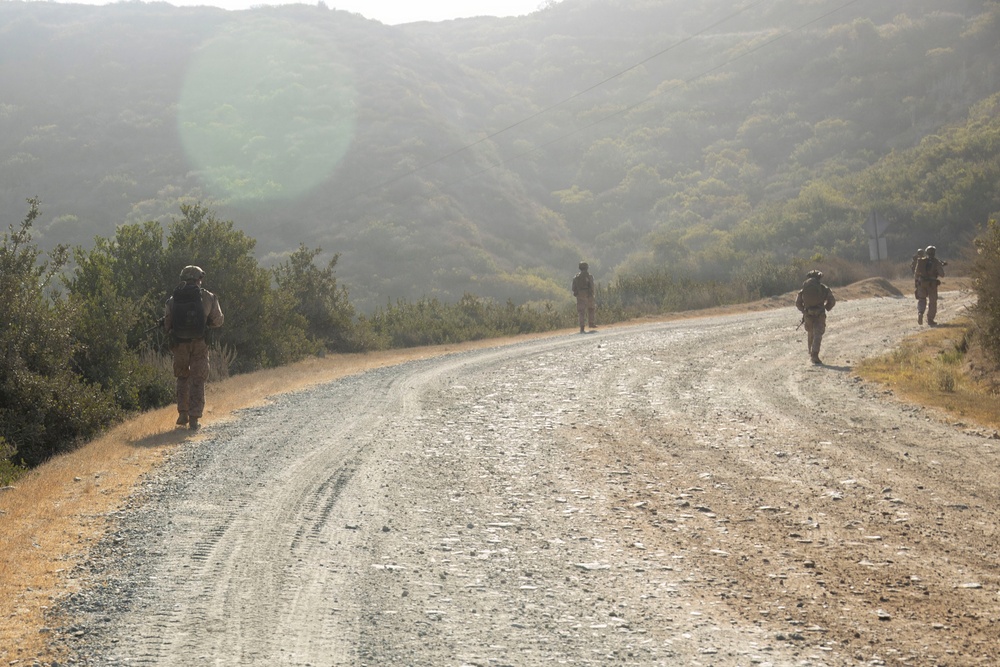 Four military personnel in full combat gear are walking along a winding gravel road through hilly, scrub-covered terrain. The soldiers are spread out in a loose patrol formation, moving away from the camera. The scene appears to be a training exercise or patrol in a dry, Mediterranean-type landscape under hazy daylight conditions.