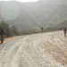 Four military personnel in full combat gear are walking along a winding gravel road through hilly, scrub-covered terrain. The soldiers are spread out in a loose patrol formation, moving away from the camera. The scene appears to be a training exercise or patrol in a dry, Mediterranean-type landscape under hazy daylight conditions.