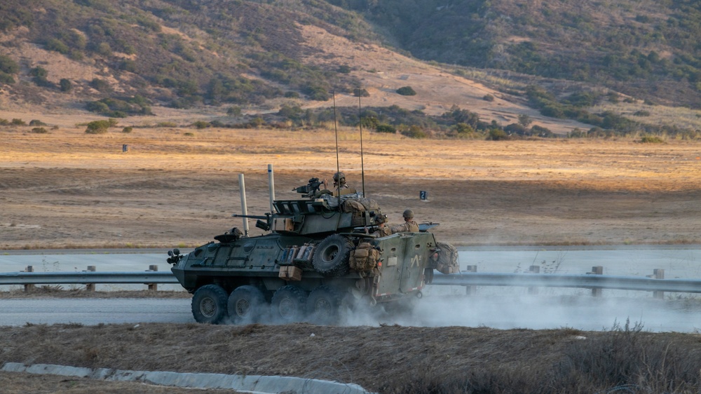 A wheeled armored fighting vehicle, consistent with an LAV-25 or similar light armored vehicle, is moving along a road or track at a military training facility. Several soldiers are visible on and around the vehicle, which is kicking up dust as it maneuvers. The terrain and landscape suggest a training environment, likely in a semi-arid region of the western United States.