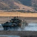 A wheeled armored fighting vehicle, consistent with an LAV-25 or similar light armored vehicle, is moving along a road or track at a military training facility. Several soldiers are visible on and around the vehicle, which is kicking up dust as it maneuvers. The terrain and landscape suggest a training environment, likely in a semi-arid region of the western United States.
