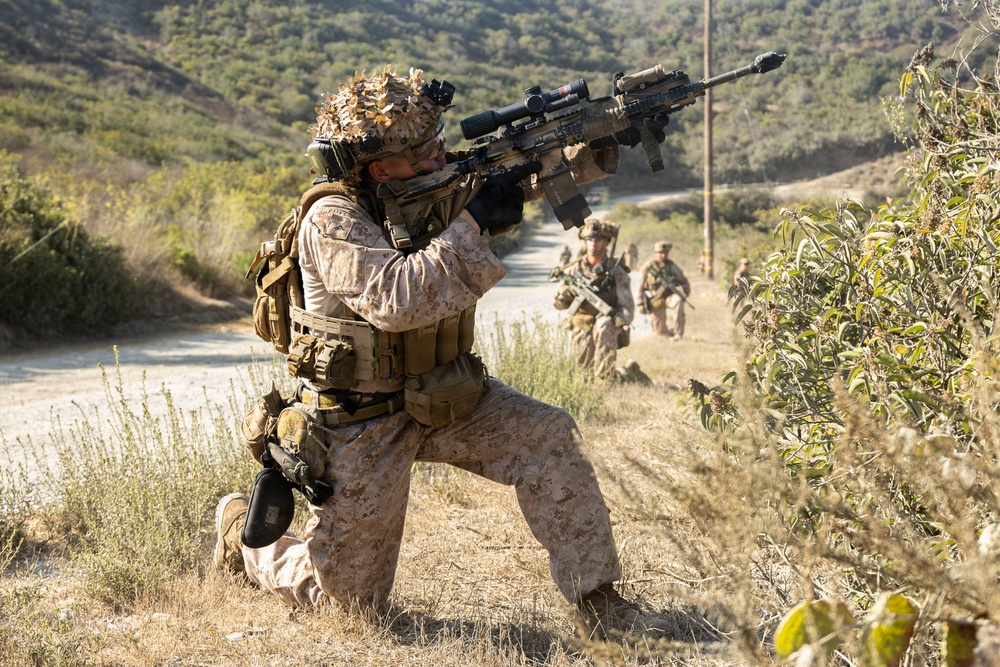 A military patrol is moving along a dirt road in a hilly, semi-arid terrain. The lead soldier is kneeling in a security position, aiming a scoped rifle toward an unseen target while teammates advance behind him. The scene appears to depict a training exercise based on the controlled environment and visible infrastructure.