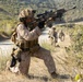 A military patrol is moving along a dirt road in a hilly, semi-arid terrain. The lead soldier is kneeling in a security position, aiming a scoped rifle toward an unseen target while teammates advance behind him. The scene appears to depict a training exercise based on the controlled environment and visible infrastructure.