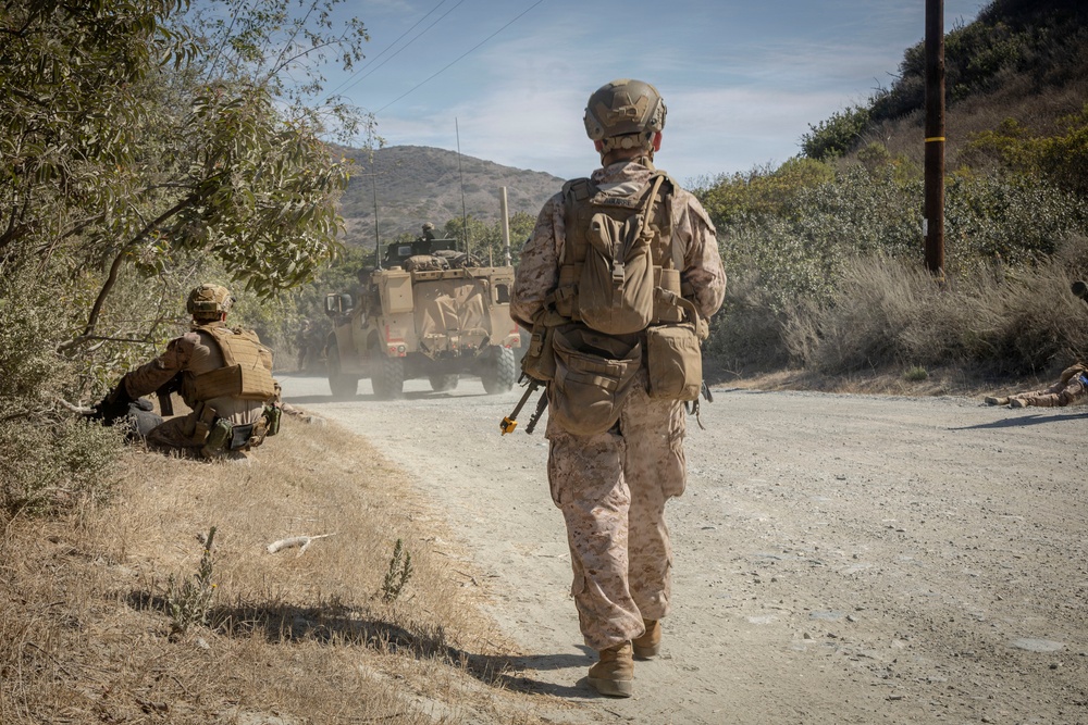 The image shows military personnel in desert camouflage uniforms operating along a dirt road in a dry, hilly terrain. A standing soldier with a backpack and tactical gear walks along the road while another soldier is positioned in a crouching stance near vegetation. Military vehicles, likely MRAPs or similar tactical trucks, are visible ahead on the road kicking up dust.