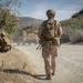 The image shows military personnel in desert camouflage uniforms operating along a dirt road in a dry, hilly terrain. A standing soldier with a backpack and tactical gear walks along the road while another soldier is positioned in a crouching stance near vegetation. Military vehicles, likely MRAPs or similar tactical trucks, are visible ahead on the road kicking up dust.