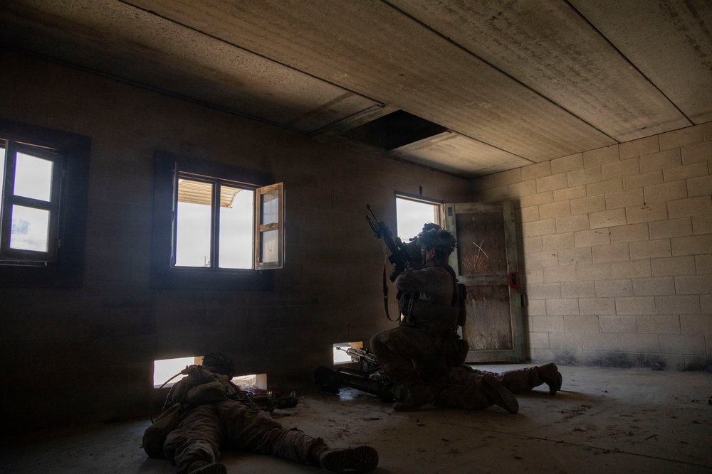The image shows two military personnel in full combat gear inside a concrete block building. One soldier is positioned seated near a doorway with a weapon raised, while another is prone on the floor near a low opening or breach point. The setting appears to be a military training facility or urban operations environment.