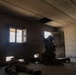 The image shows two military personnel in full combat gear inside a concrete block building. One soldier is positioned seated near a doorway with a weapon raised, while another is prone on the floor near a low opening or breach point. The setting appears to be a military training facility or urban operations environment.