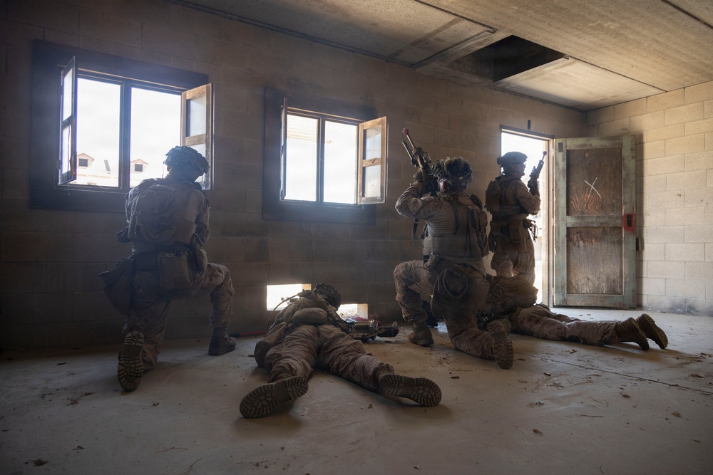 The image depicts what appears to be a military training exercise or tactical drill inside a bare concrete structure. Several soldiers in full combat gear and camouflage uniforms are positioned throughout the room, with some taking cover near windows and a doorway while others are prone on the floor. The setting resembles a military urban warfare training facility (MOUT site).