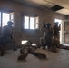 The image depicts what appears to be a military training exercise or tactical drill inside a bare concrete structure. Several soldiers in full combat gear and camouflage uniforms are positioned throughout the room, with some taking cover near windows and a doorway while others are prone on the floor. The setting resembles a military urban warfare training facility (MOUT site).