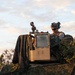 A military service member is positioned atop or behind a tan/desert-colored military vehicle, partially concealed by vegetation and a camouflage drape. The soldier is wearing full combat gear including a helmet with mounted accessories, eye protection, and body armor. The image is taken during golden hour lighting conditions, suggesting either dawn or dusk.