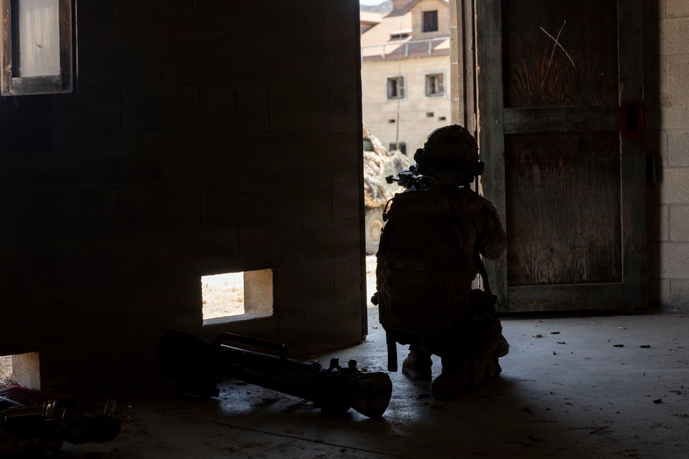A heavily equipped soldier or operator is kneeling in a low-profile position at a doorway, appearing to surveil or provide overwatch of an exterior area. The figure is silhouetted against bright exterior light, with what appears to be a rocket launcher or similar tube-launched weapon system lying on the floor nearby. The setting appears to be a damaged or austere building interior, with rubble visible outside.