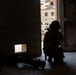 A heavily equipped soldier or operator is kneeling in a low-profile position at a doorway, appearing to surveil or provide overwatch of an exterior area. The figure is silhouetted against bright exterior light, with what appears to be a rocket launcher or similar tube-launched weapon system lying on the floor nearby. The setting appears to be a damaged or austere building interior, with rubble visible outside.