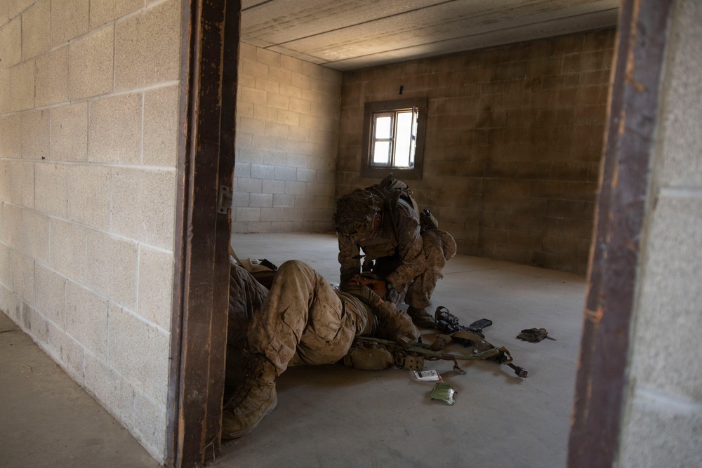 Two soldiers in full combat gear are inside a bare concrete block building, with one soldier kneeling over another who is lying on the floor. Medical supplies and equipment are scattered on the ground nearby, suggesting a tactical combat casualty care (TCCC) scenario. The setting appears to be a military training facility based on the clean, unfinished construction of the structure.