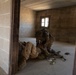 Two soldiers in full combat gear are inside a bare concrete block building, with one soldier kneeling over another who is lying on the floor. Medical supplies and equipment are scattered on the ground nearby, suggesting a tactical combat casualty care (TCCC) scenario. The setting appears to be a military training facility based on the clean, unfinished construction of the structure.