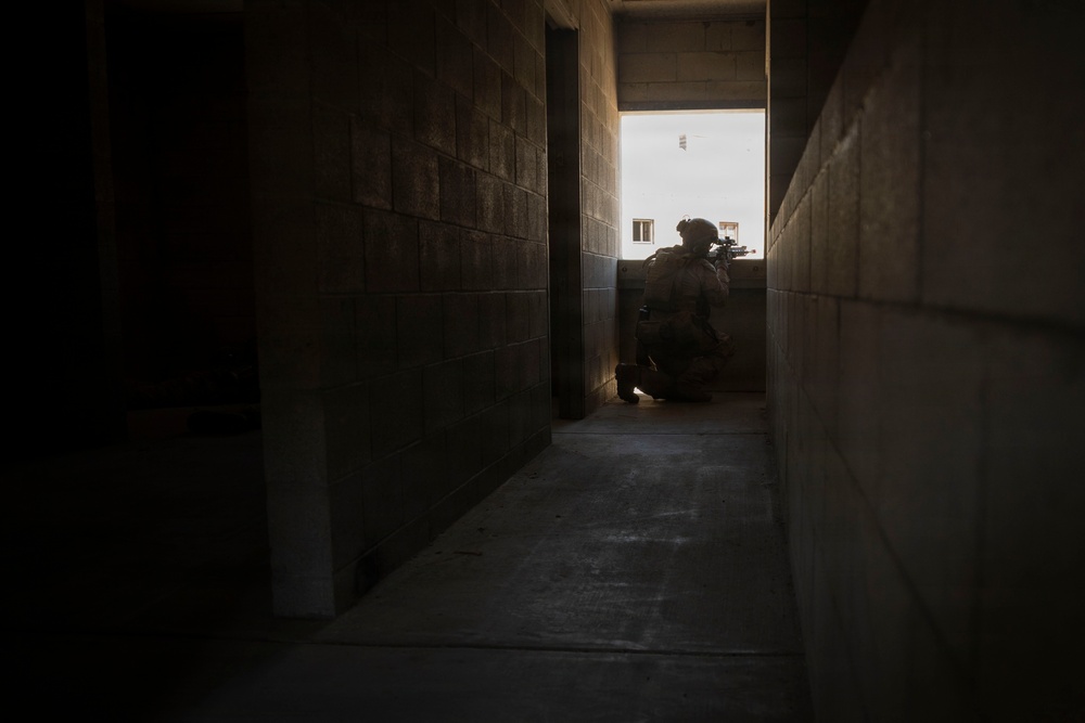 A silhouetted armed individual in tactical gear and helmet is crouched in a kneeling position at the end of a narrow concrete block corridor, aiming a weapon toward a brightly lit exterior opening. The high-contrast backlighting creates a strong silhouette effect, obscuring fine details of the figure. The setting appears to be an unfinished or austere concrete structure, consistent with a military training facility or operational environment.