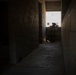 A silhouetted armed individual in tactical gear and helmet is crouched in a kneeling position at the end of a narrow concrete block corridor, aiming a weapon toward a brightly lit exterior opening. The high-contrast backlighting creates a strong silhouette effect, obscuring fine details of the figure. The setting appears to be an unfinished or austere concrete structure, consistent with a military training facility or operational environment.