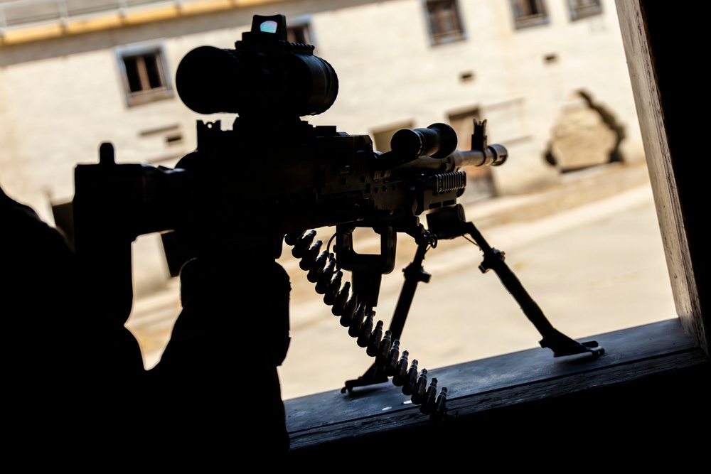 A silhouetted machine gun is positioned on a bipod mount at a window opening, with an ammunition belt visible feeding into the weapon. The scene is backlit by daylight revealing a courtyard and building exterior. The image appears to depict a defensive firing position.