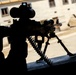 A silhouetted machine gun is positioned on a bipod mount at a window opening, with an ammunition belt visible feeding into the weapon. The scene is backlit by daylight revealing a courtyard and building exterior. The image appears to depict a defensive firing position.