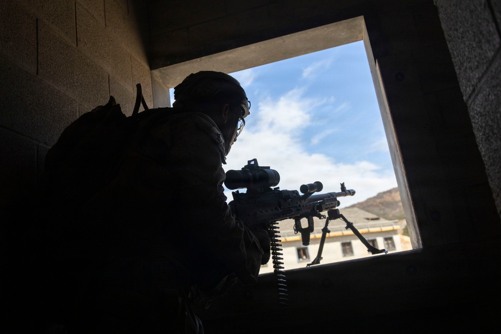 A silhouetted soldier is positioned at a window opening inside a concrete structure, aiming a belt-fed machine gun outward. The image is taken from behind and below the operator, creating a dramatic backlit composition with a blue sky and mountainous terrain visible through the aperture. A second weapon on a bipod is visible resting on the window ledge in the background.
