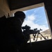 A silhouetted soldier is positioned at a window opening inside a concrete structure, aiming a belt-fed machine gun outward. The image is taken from behind and below the operator, creating a dramatic backlit composition with a blue sky and mountainous terrain visible through the aperture. A second weapon on a bipod is visible resting on the window ledge in the background.