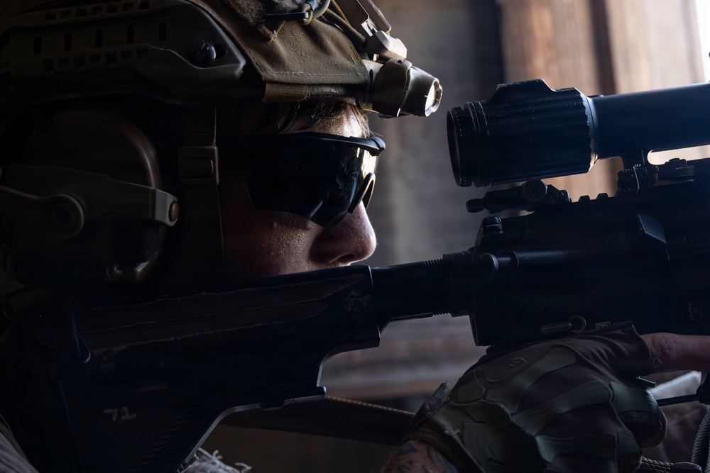 A close-up photograph of a military soldier in full tactical gear aiming down the sights of an assault rifle. The soldier is equipped with a combat helmet fitted with night vision device mounts, ballistic sunglasses, and multicam/camouflage gloves. The image is dramatically lit with strong contrast, emphasizing the soldier's focused posture.