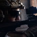 A close-up photograph of a military soldier in full tactical gear aiming down the sights of an assault rifle. The soldier is equipped with a combat helmet fitted with night vision device mounts, ballistic sunglasses, and multicam/camouflage gloves. The image is dramatically lit with strong contrast, emphasizing the soldier's focused posture.