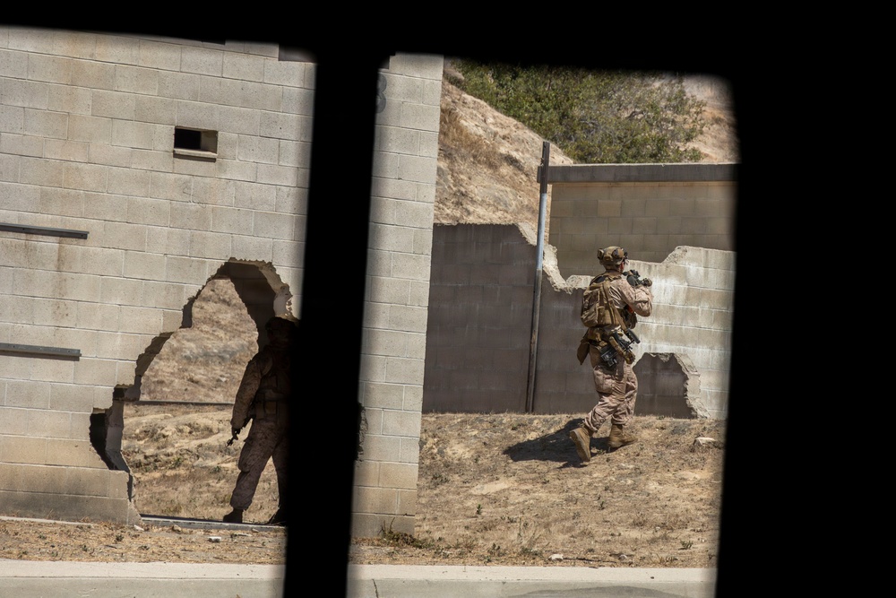 The image shows military personnel conducting what appears to be a training exercise at a mock urban combat facility. Two soldiers in desert camouflage uniforms are navigating between concrete block structures, one of which has a breached hole in the wall. The scene is photographed through a narrow opening or window frame, creating a surveillance-style perspective.