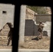 The image shows military personnel conducting what appears to be a training exercise at a mock urban combat facility. Two soldiers in desert camouflage uniforms are navigating between concrete block structures, one of which has a breached hole in the wall. The scene is photographed through a narrow opening or window frame, creating a surveillance-style perspective.