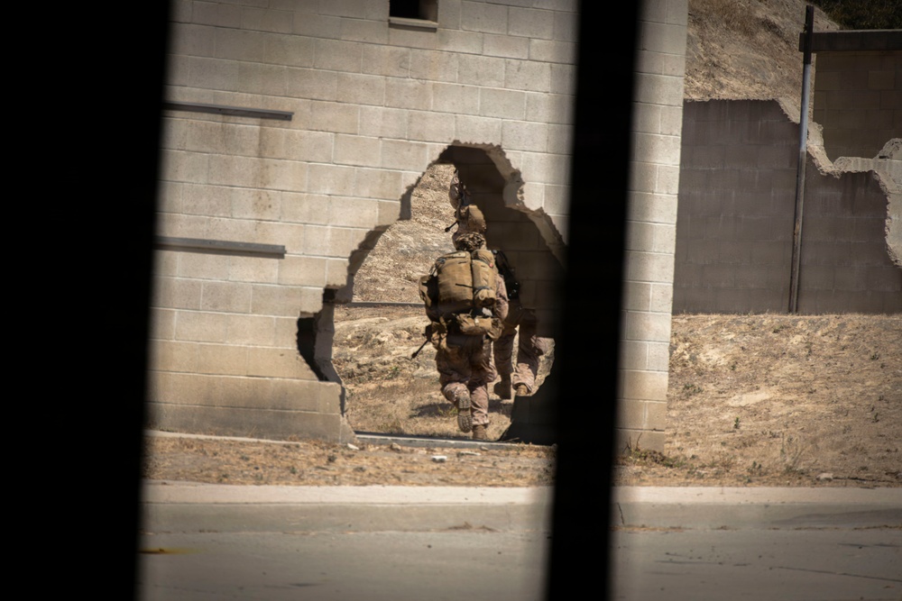 The image shows one or two military personnel in full combat gear moving through a breach in a concrete block wall. The photo is taken from a distance through what appears to be a window or vehicle opening, creating dark framing elements on the left and right sides. The setting appears to be a training facility or urban operations environment with dry, arid terrain.