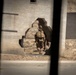 The image shows one or two military personnel in full combat gear moving through a breach in a concrete block wall. The photo is taken from a distance through what appears to be a window or vehicle opening, creating dark framing elements on the left and right sides. The setting appears to be a training facility or urban operations environment with dry, arid terrain.