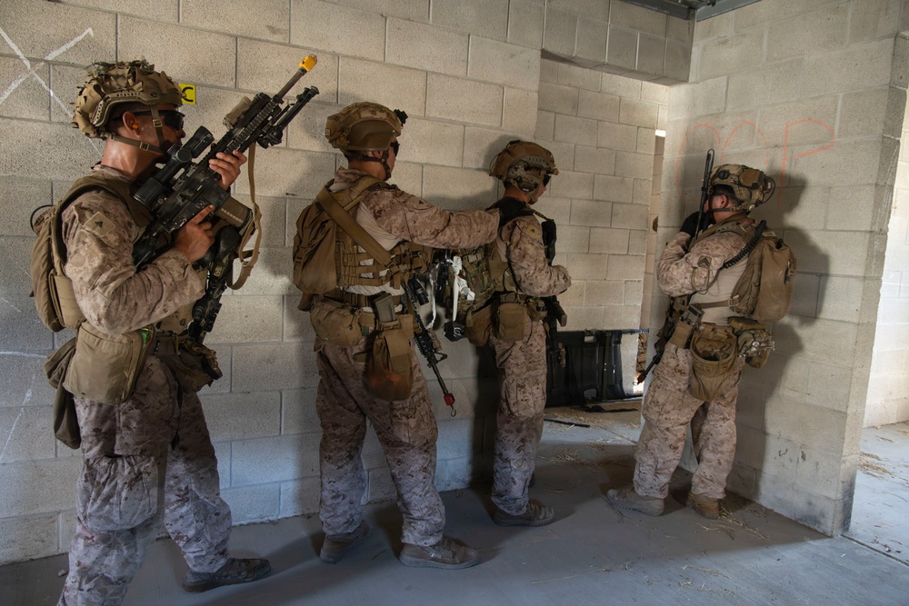 Four military personnel in desert camouflage uniforms are conducting what appears to be a military training exercise inside a concrete block building. They are equipped with full tactical gear, helmets, body armor, and weapons, and appear to be preparing to breach or clear a room. The setting is an urban operations training facility, indicated by markings on the walls and the controlled environment.