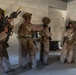 Four military personnel in desert camouflage uniforms are conducting what appears to be a military training exercise inside a concrete block building. They are equipped with full tactical gear, helmets, body armor, and weapons, and appear to be preparing to breach or clear a room. The setting is an urban operations training facility, indicated by markings on the walls and the controlled environment.