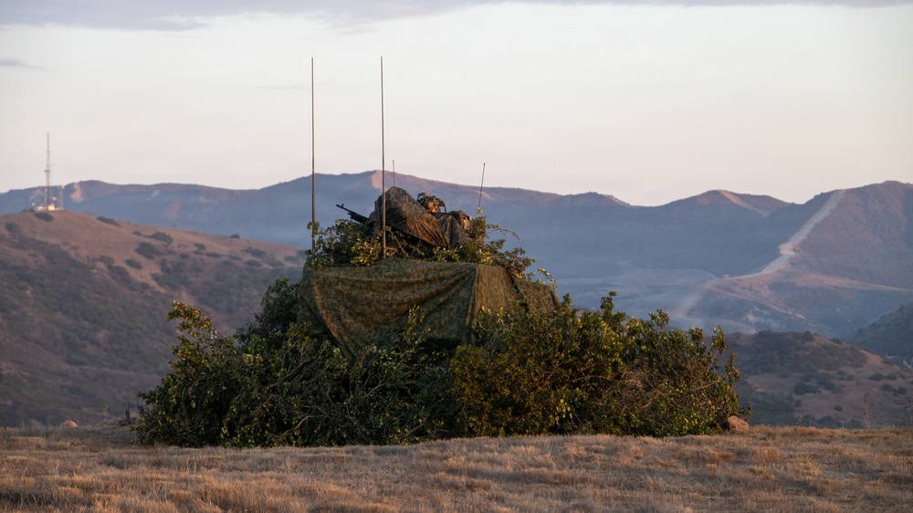 A military position concealed with camouflage netting and natural vegetation (brush/shrubs) on an open hillside. At least one armed soldier is visible atop the concealed position, which also features multiple radio antennas. The setting appears to be a training range or operational area in a dry, mountainous landscape at dusk or dawn.