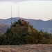 A military position concealed with camouflage netting and natural vegetation (brush/shrubs) on an open hillside. At least one armed soldier is visible atop the concealed position, which also features multiple radio antennas. The setting appears to be a training range or operational area in a dry, mountainous landscape at dusk or dawn.