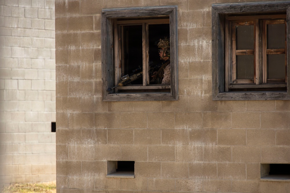 A military service member in full combat gear and helmet is positioned at an open window of a concrete block building, holding a rifle in a surveillance or defensive posture. The building appears to be a training facility or mock urban environment, constructed from cinder blocks with weathered wooden window frames. The scene is consistent with military urban warfare training exercises.