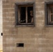 A military service member in full combat gear and helmet is positioned at an open window of a concrete block building, holding a rifle in a surveillance or defensive posture. The building appears to be a training facility or mock urban environment, constructed from cinder blocks with weathered wooden window frames. The scene is consistent with military urban warfare training exercises.