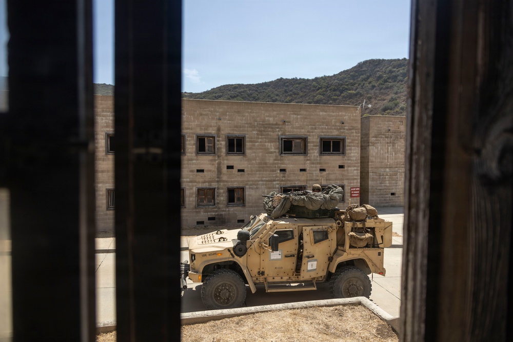 The image is taken through a doorway or window frame with vertical bars, looking out onto a courtyard area where a tan/desert-colored military tactical vehicle (JLTV or HMMWV variant) is parked. A soldier appears to be positioned on top of the vehicle, possibly manning a weapon system or observing. The setting appears to be a military training facility with concrete block buildings and a hillside visible in the background.