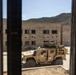 The image is taken through a doorway or window frame with vertical bars, looking out onto a courtyard area where a tan/desert-colored military tactical vehicle (JLTV or HMMWV variant) is parked. A soldier appears to be positioned on top of the vehicle, possibly manning a weapon system or observing. The setting appears to be a military training facility with concrete block buildings and a hillside visible in the background.