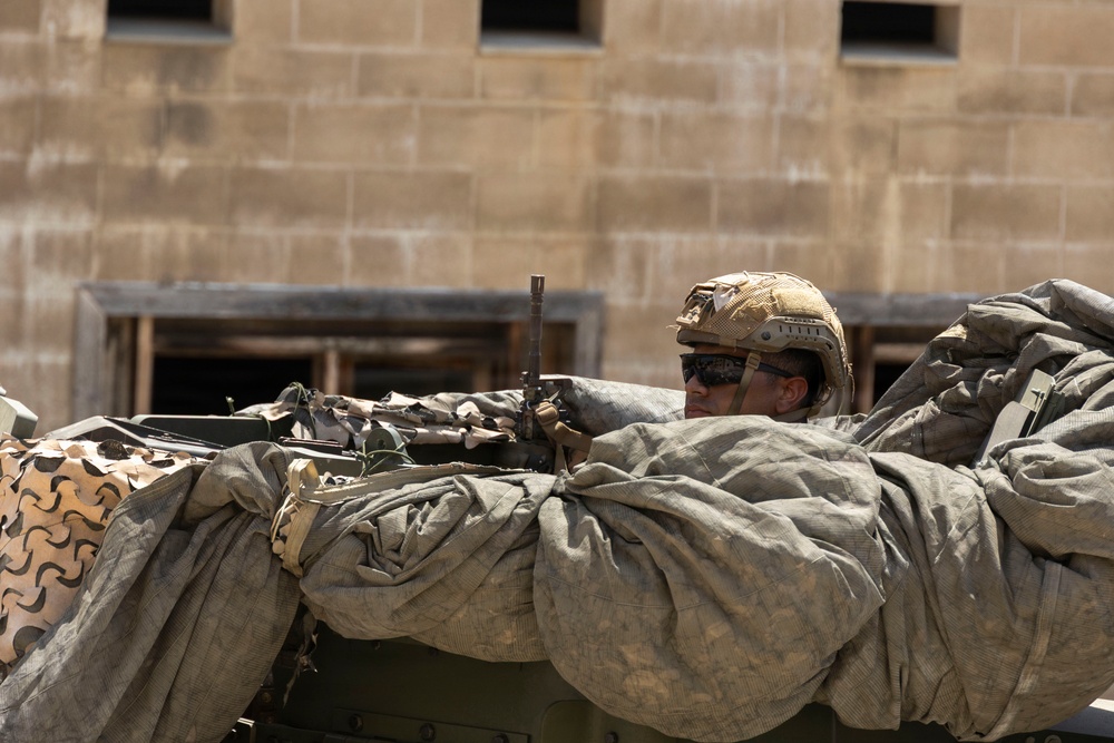 A soldier in full combat gear is positioned behind a military vehicle loaded with equipment and camouflage netting. The soldier is aiming a rifle while taking cover, suggesting a tactical or training operation. The background shows a tan concrete block building with rectangular window openings.