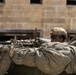 A soldier in full combat gear is positioned behind a military vehicle loaded with equipment and camouflage netting. The soldier is aiming a rifle while taking cover, suggesting a tactical or training operation. The background shows a tan concrete block building with rectangular window openings.