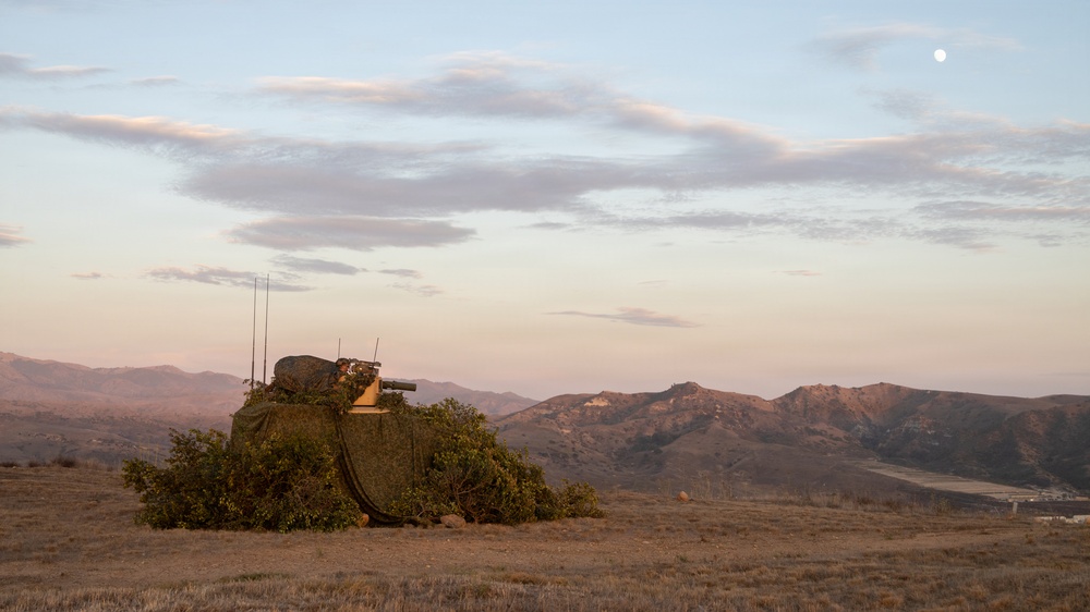 A military vehicle or weapons system is heavily camouflaged with netting and natural vegetation on an open, arid hilltop at dusk or dawn. The equipment appears to include a mounted weapon or optical system along with multiple radio antennas. The scene is set against a backdrop of rolling mountains with a visible moon in the partially cloudy sky.